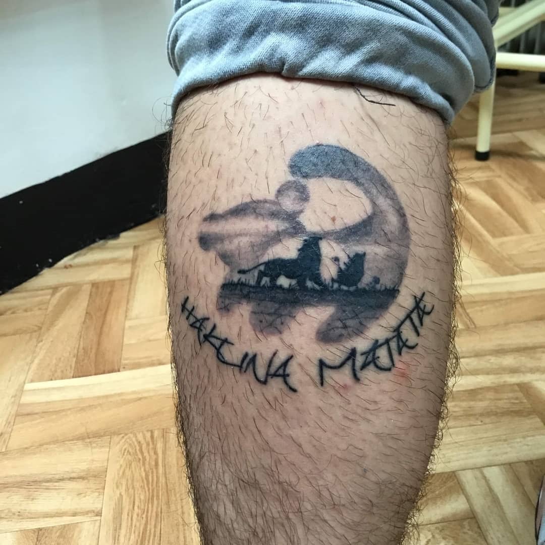 Tattoo of a scene with a lion and cub, with text "Hakuna Matata," on a leg